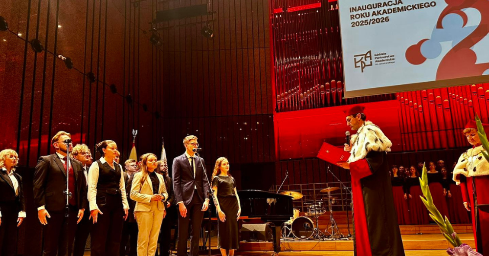 W dużej sali z podestem i organami w tle odbywa się uroczystość inauguracji roku akademickiego. Po prawej stronie stoi dostojna postać w todze i futrzanej pelerynie, trzymająca mikrofon i czytająca przemówienie z czerwonej teczki. Po lewej, na scenie przed mikrofonami, ustawiona grupa młodych osób — studenci i studentki — w eleganckich strojach, stoją prosto i poważnie. W tle widać część chóru oraz instrumentalistów, a nad sceną – baner z napisem „Inauguracja Roku Akademickiego 2025/2026”. Sceneria jest formalna, dekorowana czerwoną poświatą i przemyślanym oświetleniem, nadając wydarzeniu podniosły charakter.
