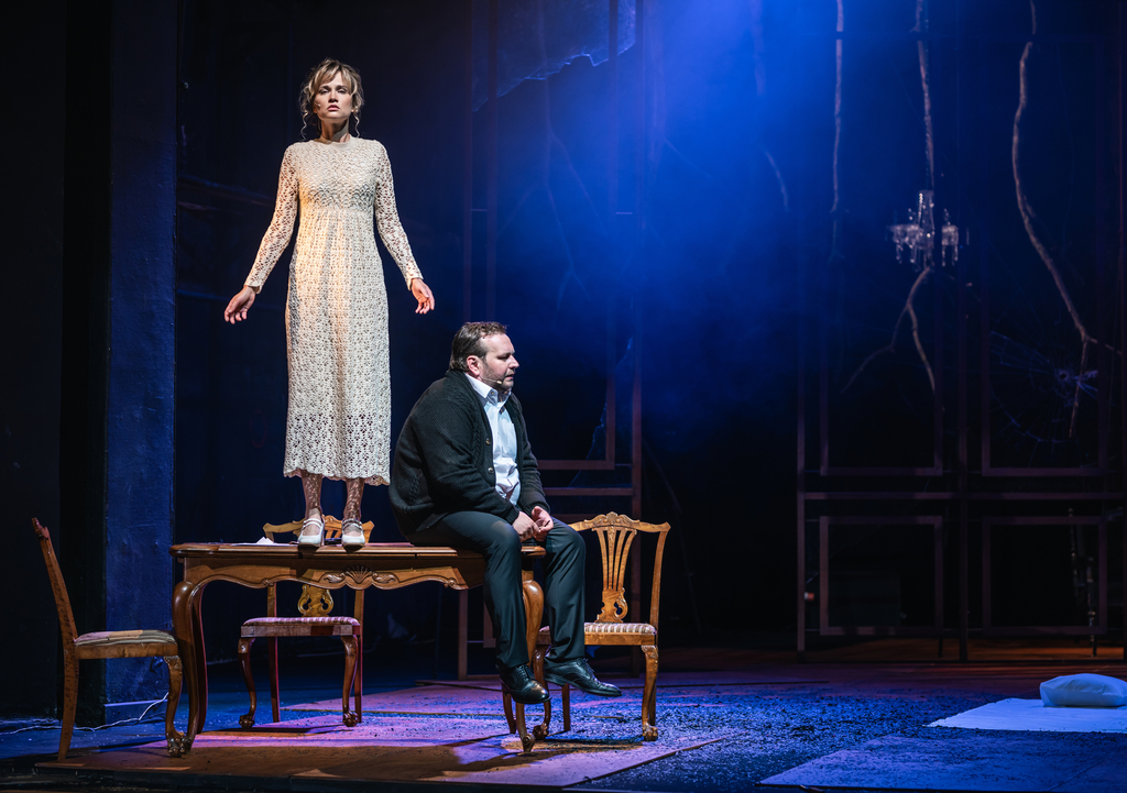 Fotos ze spektaklu. Kobieta stoi na stole, obok siedzi mężczyzna.