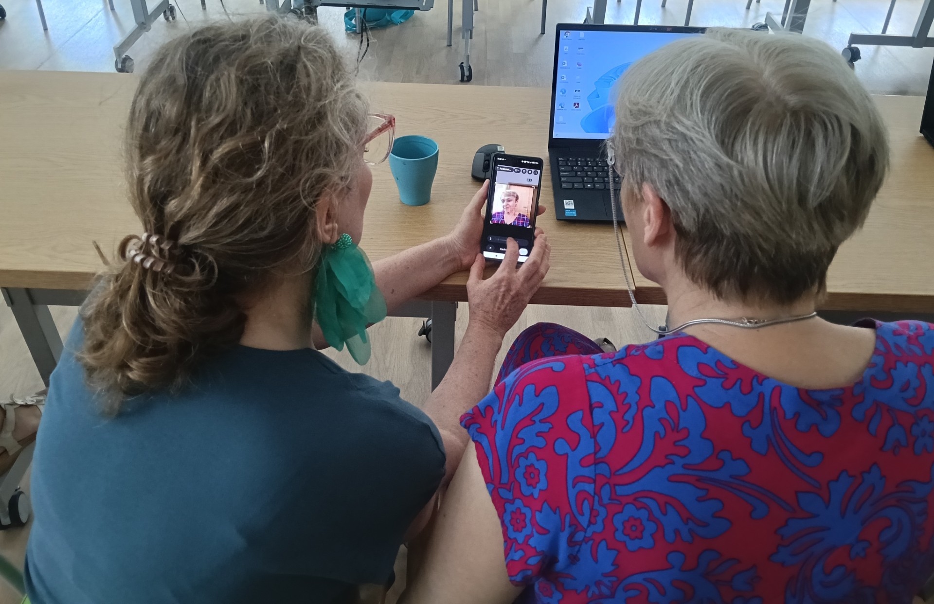 dwie kobiety siedzące tyłem do nas, wpatrują się w ekran telefonu.