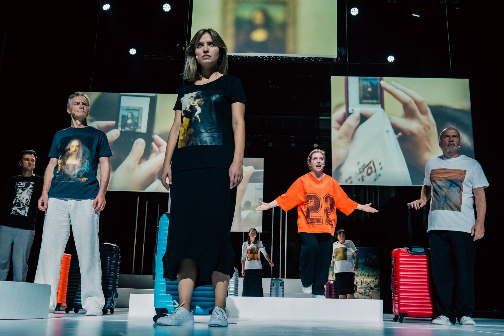 Na zdjęciu widać grupę osób stojących i patrzących w jeden punkt gdzieś w oddali. Są to mężczyźni i kobiety, ubrani jak turyści, obok każdej osoby stoi walizka. W tle widać wielkie ekrany ze zdjęciami turystów.