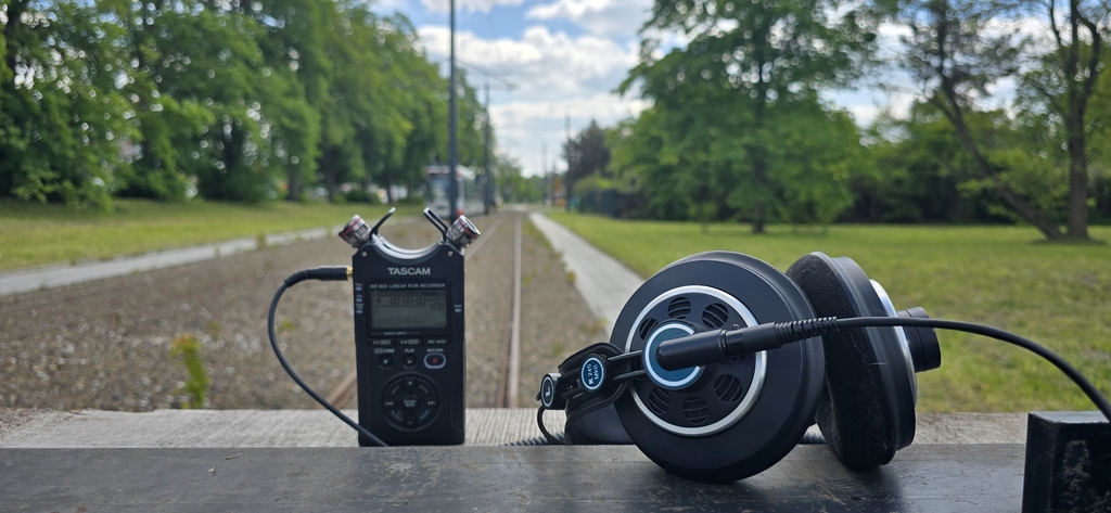 Na zdjęciu widać urządzenie nagrywające typu dyktafon w kolorze czarnym, po prawej stronie leżą słuchawki. W drugim planie o oddali widać fragment parku, zielone drzewa, fragment niebieskiego nieba.