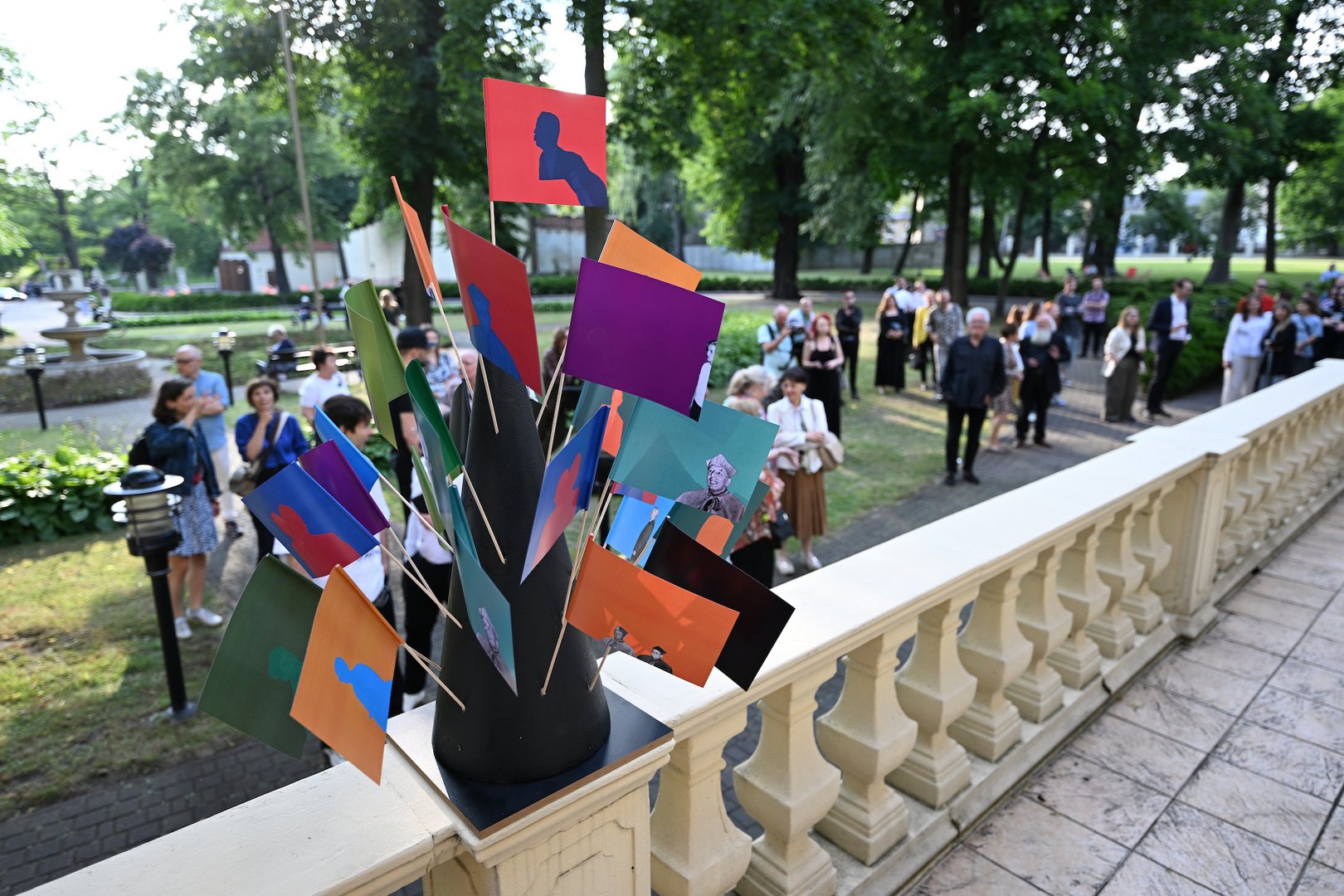 Na zdjęciu widać park, w którym jest dużo ludzi. Jest wiosna, drzewa są zielone. Na pierwszym planie widoczna balustrada z betonu na której stoi stojak z kolorowymi chorągiewkami. W tle chodzą wspomniani ludzie.