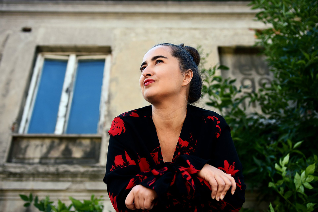Anka Leśniak, fot. Agnieszka Cytacka Na zdjęciu kobieta siedząca przed budynkiem. Patrzy w dal, ma uniesioną głowę. Ubrana w czarną bluzkę z czerwonym wzorem..