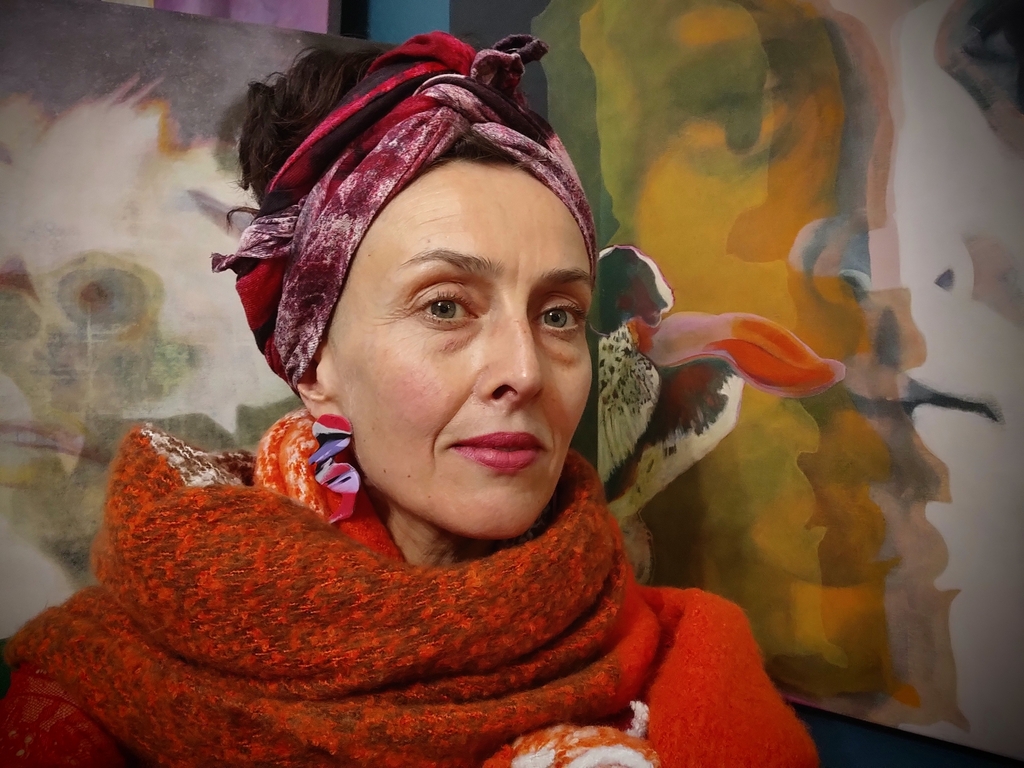 Na zdjęciu widać twarz kobiety, która patrzy w naszą stronę. Ubrana jest w sweter w kolorze pomarańczowym. Za nią widoczne są fragmenty obrazów, które wiszą na ścianie.