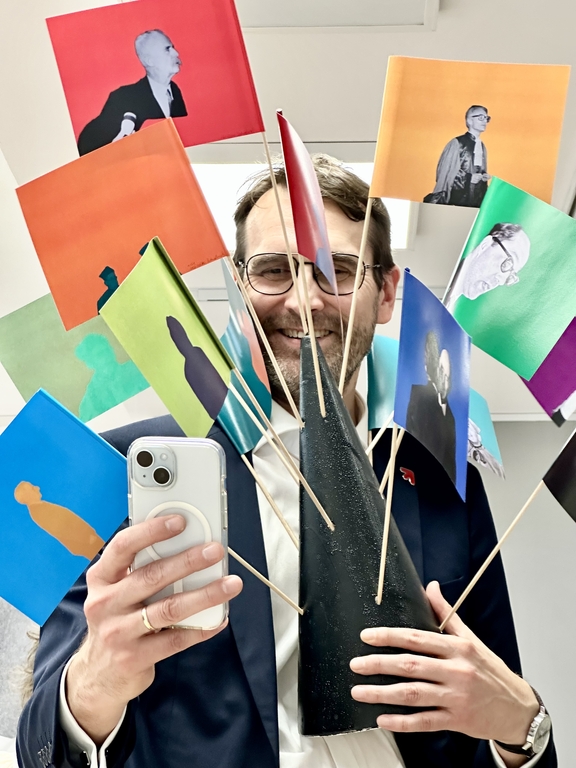 Na zdjęciu widać mężczyznę, który trzyma chorągiewki zatknięte w stożku ze styropianu. Mężczyzna trzyma w drugiej dłoni smartfon i robi nim zdjęcie.