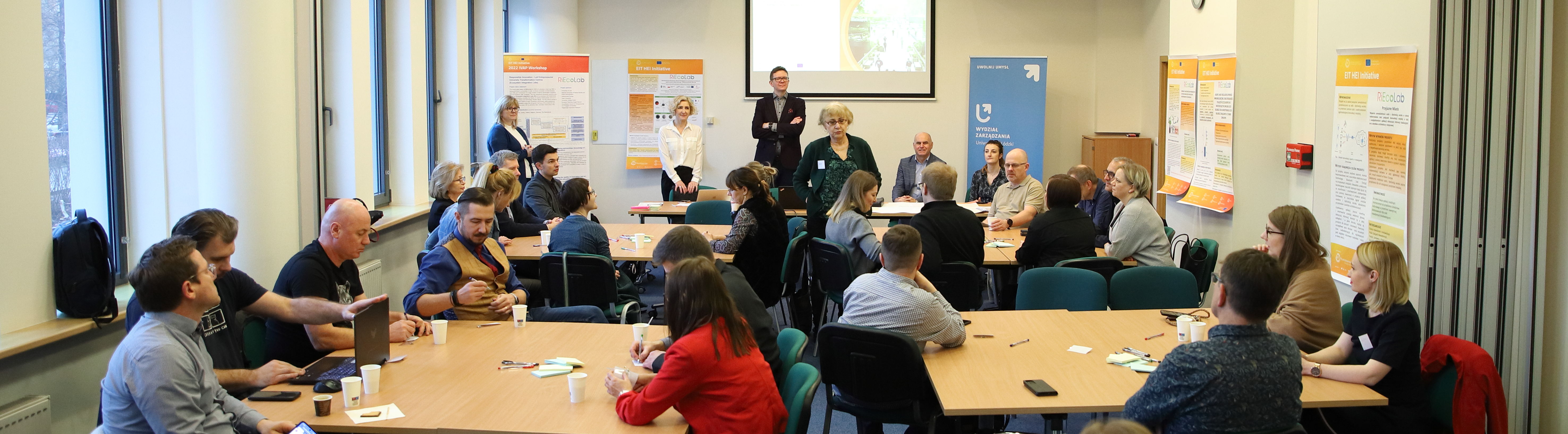 grupa osób siedzi w sali, a dwóch prowadzących omawia temat