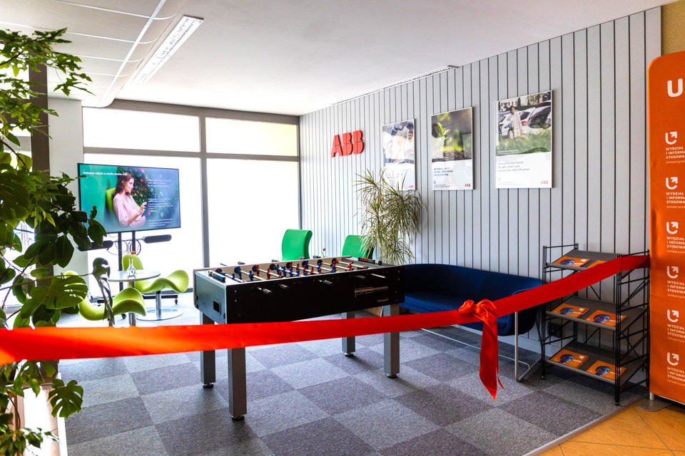 The relaxation zone with: table football, a rack with leaflets, 3 pictures in a clip frame, the ABB logo (red capital letters ABB ), backrests of green armchairs, a TV set, a piece of a green flower and a red sash that separates the zone
