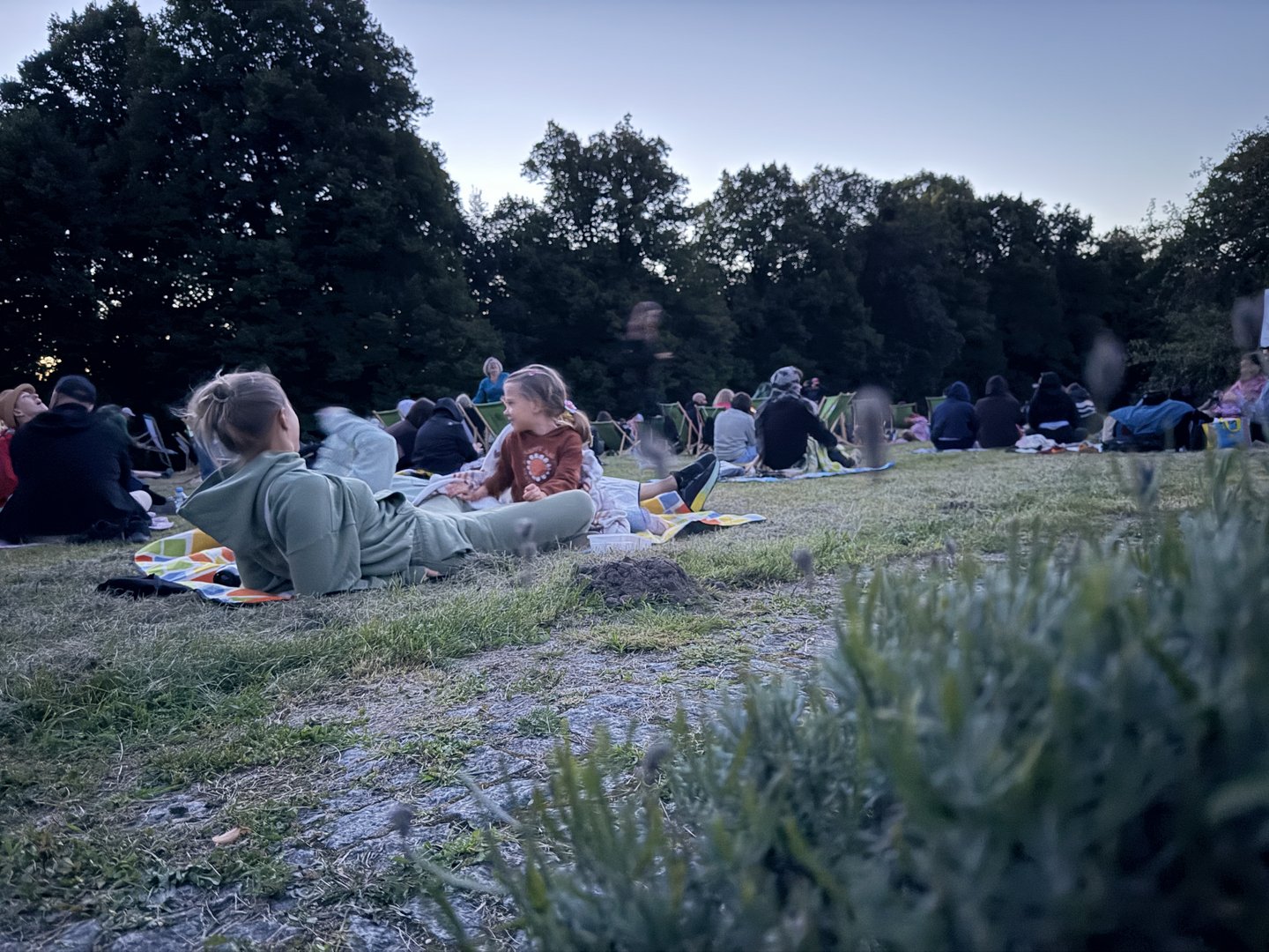 Kobieta i dziecko siedzą na kocu na trawie podczas wydarzenia plenerowego. W tle widoczni są inni uczestnicy oraz drzewa.