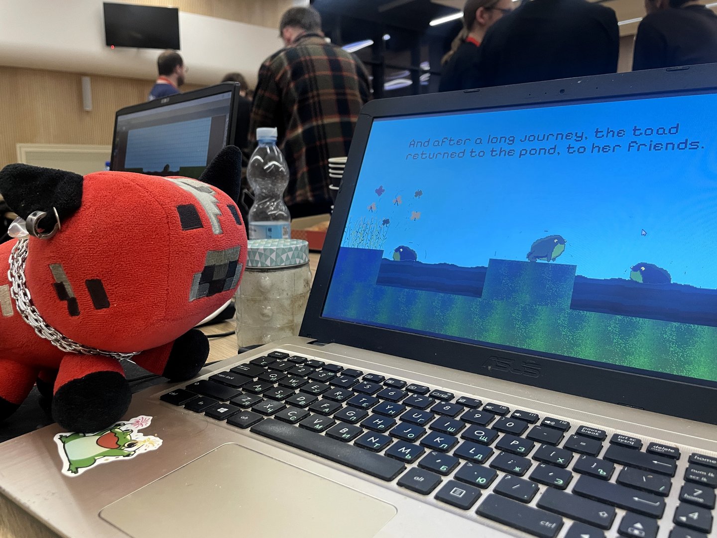 Na zdjęciu widać otwarty laptop stojący na stole podczas wydarzenia. Na ekranie wyświetlana jest gra komputerowa w stylu pikselowym z żabą na tle nieba i wody oraz tekstem końcowym gry. Obok laptopa leży czerwono-czarna pluszowa maskotka w kształcie zwierzątka z kolczykiem i łańcuszkiem. Na stole znajdują się także butelka wody i słoik. W tle widać grupę osób stojących w sali, co sugeruje, że zdjęcie zostało wykonane podczas maratonu tworzenia gier.
