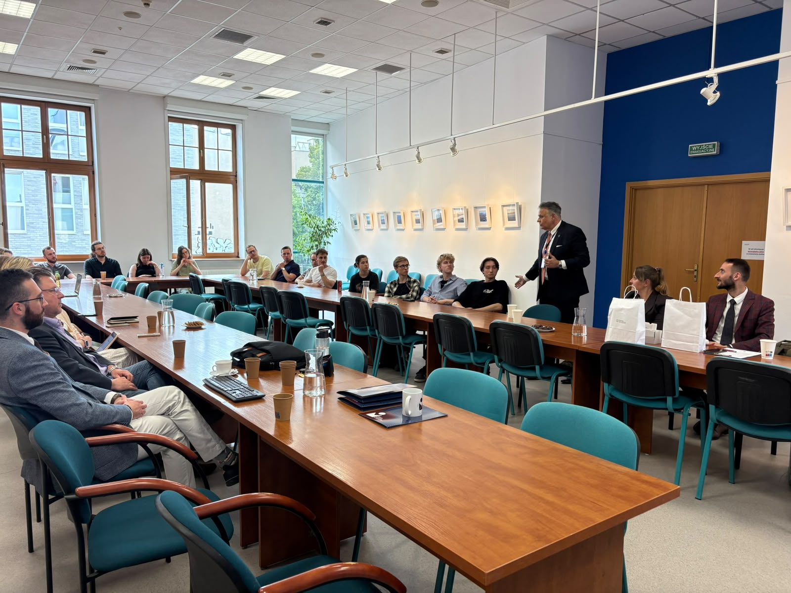 Szerokokątne ujęcie jasnej sali konferencyjnej z ustawionymi w podkowę stołami i zielonymi krzesłami; przy stołach siedzi grupa uczestników, a jedna osoba stojąca przemawia do zgromadzonych. Na stołach znajdują się dzbanki z wodą, kubki, notatniki i przekąski. W tle widoczne są duże okna, roślina doniczkowa oraz rząd oprawionych zdjęć na ścianie, a także drzwi z oznaczeniem „Wyjście ewakuacyjne”. Pomieszczenie ma sufit podwieszany z oświetleniem i projektorem. Szerokokątne ujęcie jasnej sali konferencyjnej z ustawionymi w podkowę stołami i zielonymi krzesłami; przy stołach siedzi grupa uczestników, a jedna osoba stojąca przemawia do zgromadzonych. Na stołach znajdują się dzbanki z wodą, kubki, notatniki i przekąski. W tle widoczne są duże okna, roślina doniczkowa oraz rząd oprawionych zdjęć na ścianie, a także drzwi z oznaczeniem „Wyjście ewakuacyjne”. Pomieszczenie ma sufit podwieszany z oświetleniem i projektorem.