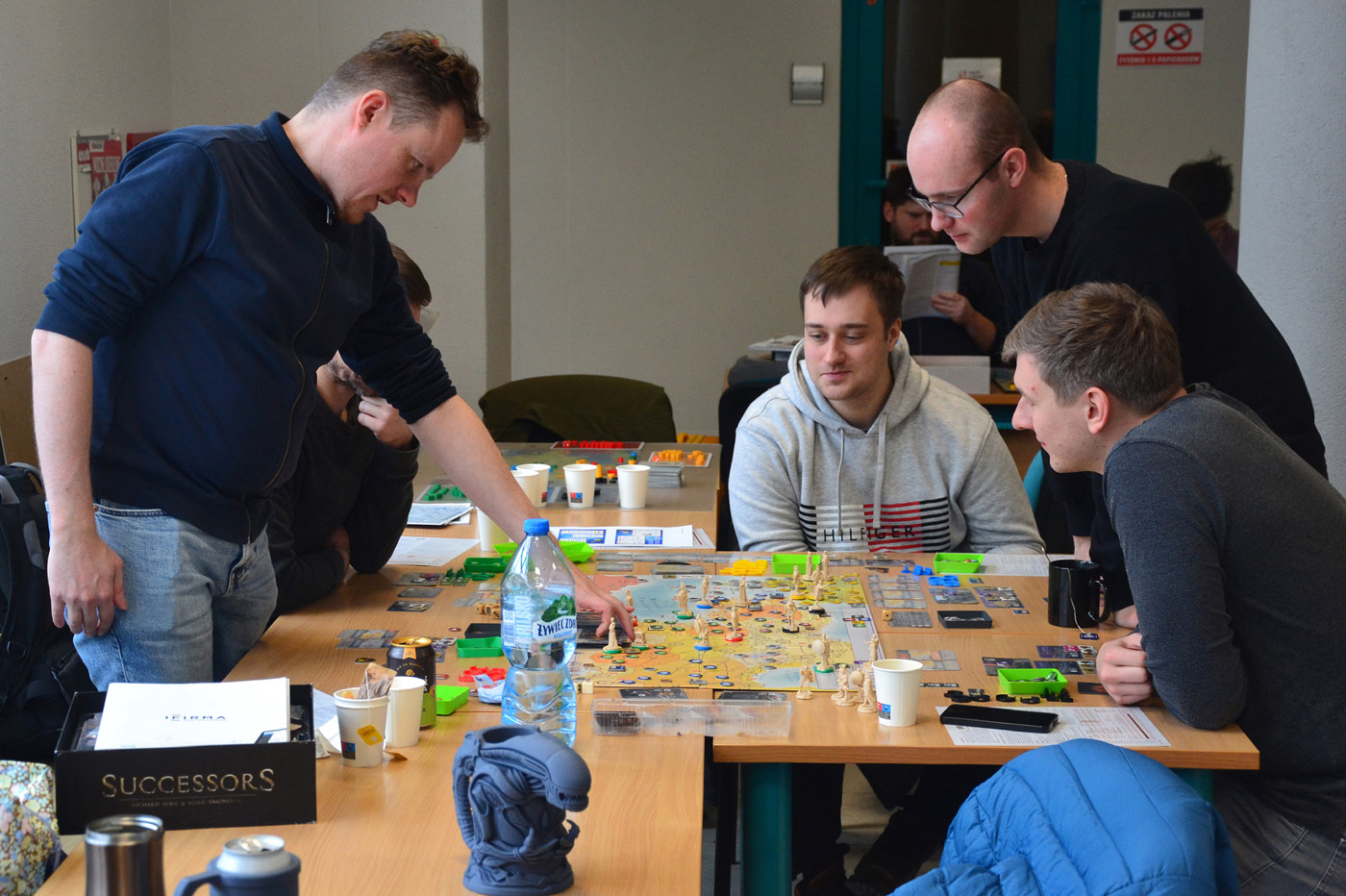Pięć osób grających w zaawansowaną grę planszową „Successors” przy stole w przestrzeni wspólnej. Na stole widoczne są plansza, figurki, karty, żetony oraz napoje i przekąski. Jedna osoba wskazuje na planszę, pozostali uważnie obserwują lub dyskutują. W tle kolejna osoba czyta instrukcję, co sugeruje atmosferę nauki i zaangażowania.