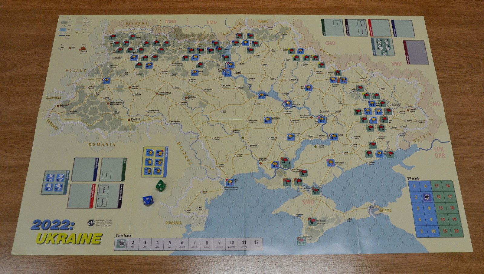 Plansza strategicznej gry wojennej „2022: UKRAINE” przedstawiająca Ukrainę i sąsiednie kraje w układzie heksagonalnym. Na mapie widoczne są znaczniki jednostek w różnych kolorach, oznaczenia miast takich jak Kijów, Charków, Odessa czy Mariupol, tor punktów zwycięstwa oraz tor rund od kwietnia do grudnia. Obecne są także kostki do gry i pola organizacyjne po bokach mapy.