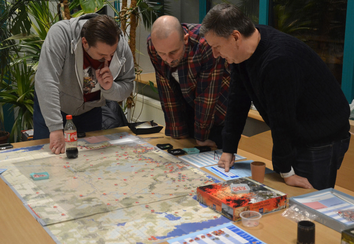 Trzy osoby grające w strategiczną grę planszową „Stalingrad: Verdun on the Volga”. Na stole rozłożona jest szczegółowa mapa z czerwonymi i niebieskimi znacznikami, karty, arkusze pomocnicze i pojemniki z elementami gry. Gracze są skupieni na analizie sytuacji, a tło wskazuje na swobodną przestrzeń klubową lub domową.