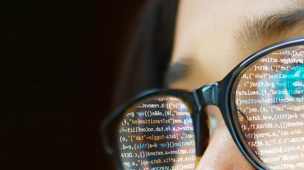 widoczna z bliska twarz i okulary w których odbija się ekran komputera z programem do kodowania