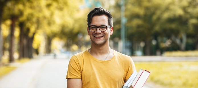 kolorowe zdjęcie przedstawiające młodego uśmiechniętego mężczyznę w okularach trzymającego w rękach kilka książek; mężczyzna stroi w parku na tle drzew