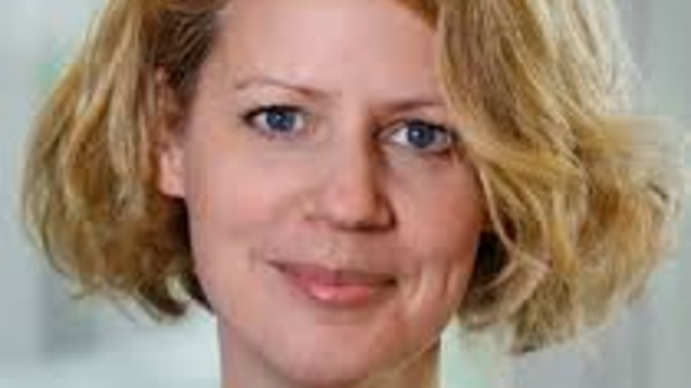 Portrait of a woman with short, wavy blonde hair wearing a light blue blouse, standing indoors against a softly blurred background and smiling gently at the camera.