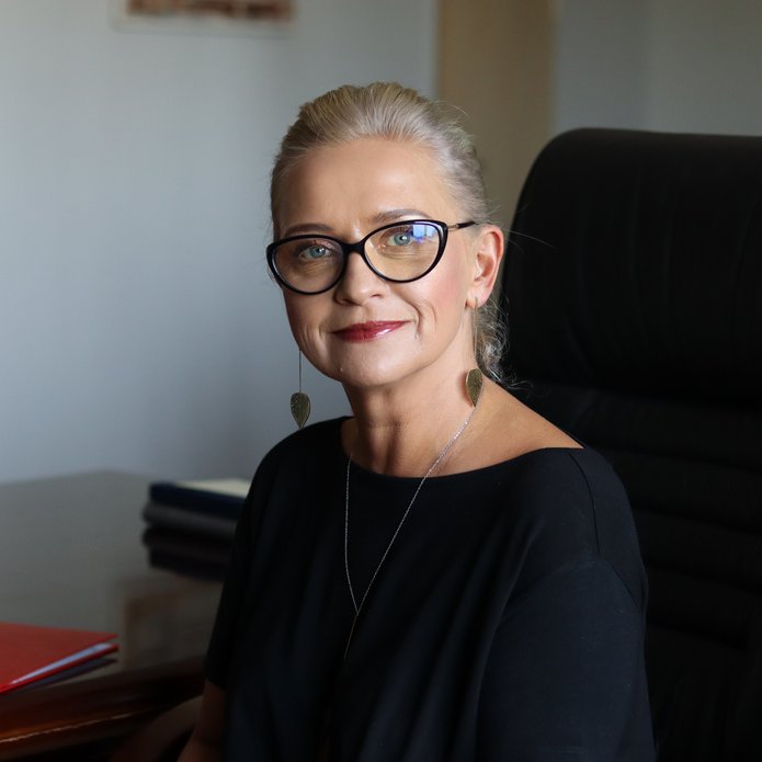 Pani Profesor o blond włosach z okularami na nosie ubrana w czarną sukienkę.