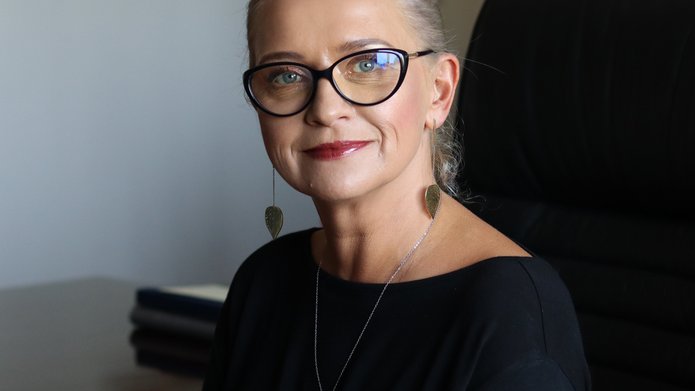 Pani Profesor o blond włosach z okularami na nosie ubrana w czarną sukienkę.