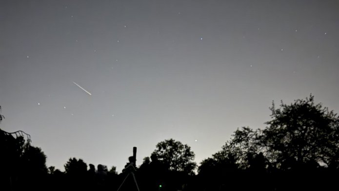 Nocne niebo nad Ogrodem Botanicznym, usiane gwiazdami. W lewej części nieba widoczna jest jasna, krótka smuga meteoru – Perseidy. Na dole zdjęcia, na ciemnej polanie, widać sylwetki osób leżących na kocach oraz teleskop ustawiony na statywie. W tle znajdują się ciemne sylwetki drzew.