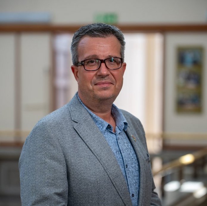 Mężczyzna stoi przodem do aparatu, ubrany w garnitur. 