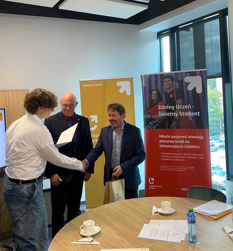 Trzy osoby w sali konferencyjnej. Dwóch mężczyzn w garniturach, stojących po prawej stronie, wręcza certyfikat młodszemu mężczyźnie po lewej stronie. Starszy z nich uśmiecha się i podaje rękę młodszemu mężczyźnie, który jest ubrany w białą koszulę i dżinsy. W tle znajdują się dwa roll-upy reklamowe: : jeden żółty z logo Wydziału Nauk o Wychowaniu, drugi czerwony z logo Zdolny uczeń-świetny student. Na stole przed nimi widać filiżanki, butelkę wody i kilka dokumentów.