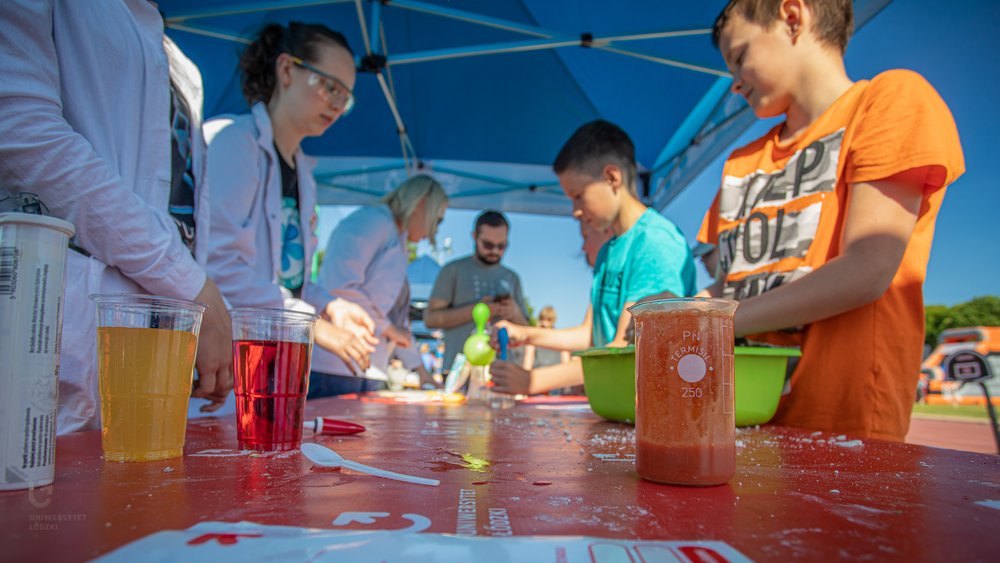 studenci w fartuchach id zieci stojące przy stole, na którym ustawione są naczynia z kolorowymi cieczami