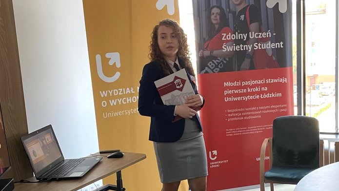 Na zdjęciu znajduje się kobieta w sali konferencyjnej. Jest ubrana w elegancką marynarkę i spódnicę, trzyma dokumenty w rękach i uśmiecha się do kamery. W tle znajdują się dwa roll-upy: jeden żółty z logo Wydziału Nauk o Wychowaniu, drugi czerwonyz logo Zdolny uczeń-świetny student. Obok kobiety znajduje się stolik z laptopem, a w tle widać duże okna, przez które wpada naturalne światło. Pomieszczenie jest jasne i nowoczesne, z krzesłami ustawionymi wokół okrągłego stołu.