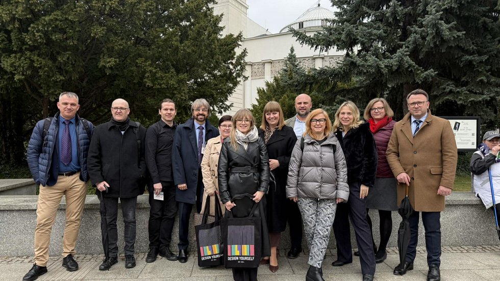 grupa ludzi stojąca przed budynkiem sejmu RP, są to kobiety i mężczyzni, stoją na chodniku, w tle widoczny budynek sali plenarnej sejmu.