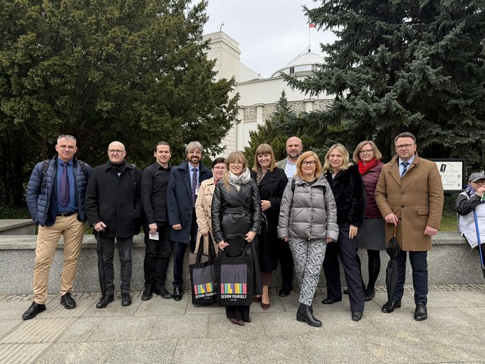 grupa ludzi stojąca przed budynkiem sejmu RP, są to kobiety i mężczyzni, stoją na chodniku, w tle widoczny budynek sali plenarnej sejmu.