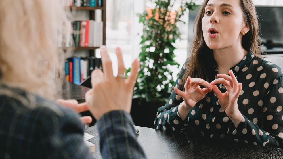 Dwie kobiety siedzące naprzeciwko siebie, uczą się języka migowego