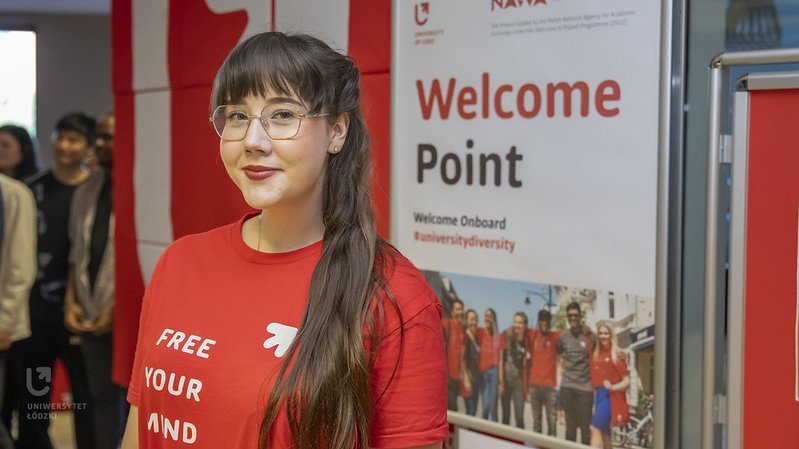 A girl in a red T-shirt of the University of Lodz standing in front of a poster with the words "Welcome Point"