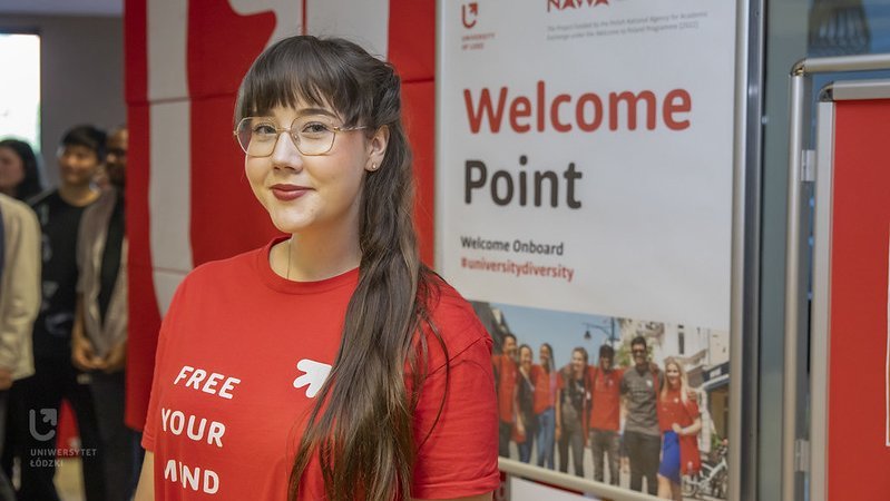 Dziewczyna w czerwonej koszulce UŁ stoi na tle plakatu z napisem "Welcome Point"