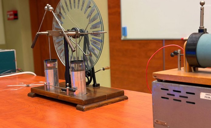 Na stole w sali laboratoryjnej znajduje się zabytkowy elektroskop z dużą okrągłą tarczą i metalowymi elementami, obok którego ustawiono inne urządzenie do doświadczeń z elektryczności. W tle widoczna jest tablica oraz wyposażenie sali dydaktycznej.