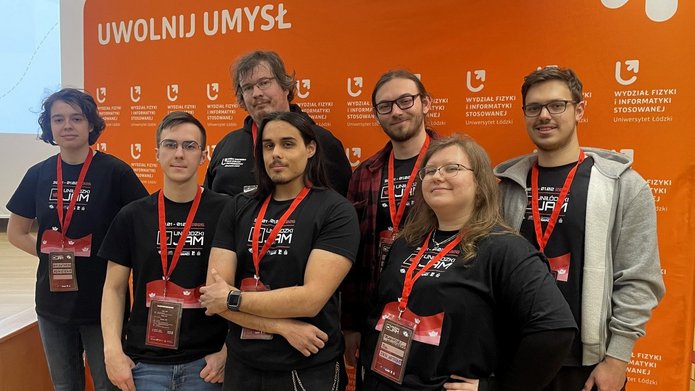 Na zdjęciu widać grupę ośmiu osób stojących obok siebie i pozujących do zdjęcia. Wszyscy mają na sobie czarne koszulki z napisem „UniŁódzki Jam 2026” oraz czerwone identyfikatory na szyjach. Stoją na tle pomarańczowej ścianki z logo Wydziału Fizyki i Informatyki Stosowanej Uniwersytetu Łódzkiego i hasłem „Uwolnij umysł”. Osoby są uśmiechnięte i wyglądają na zadowolone.