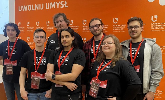 Na zdjęciu widać grupę ośmiu osób stojących obok siebie i pozujących do zdjęcia. Wszyscy mają na sobie czarne koszulki z napisem „UniŁódzki Jam 2026” oraz czerwone identyfikatory na szyjach. Stoją na tle pomarańczowej ścianki z logo Wydziału Fizyki i Informatyki Stosowanej Uniwersytetu Łódzkiego i hasłem „Uwolnij umysł”. Osoby są uśmiechnięte i wyglądają na zadowolone.