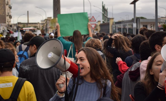 Na zdjęciu widać tłum ludzi. Na przodzie stoi dziewczyna z długimi włosami, w prawej dłoni trzyma biały megafon przez który coś krzyczy.