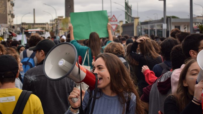 Na zdjęciu widać tłum ludzi. Na przodzie stoi dziewczyna z długimi włosami, w prawej dłoni trzyma biały megafon przez który coś krzyczy.