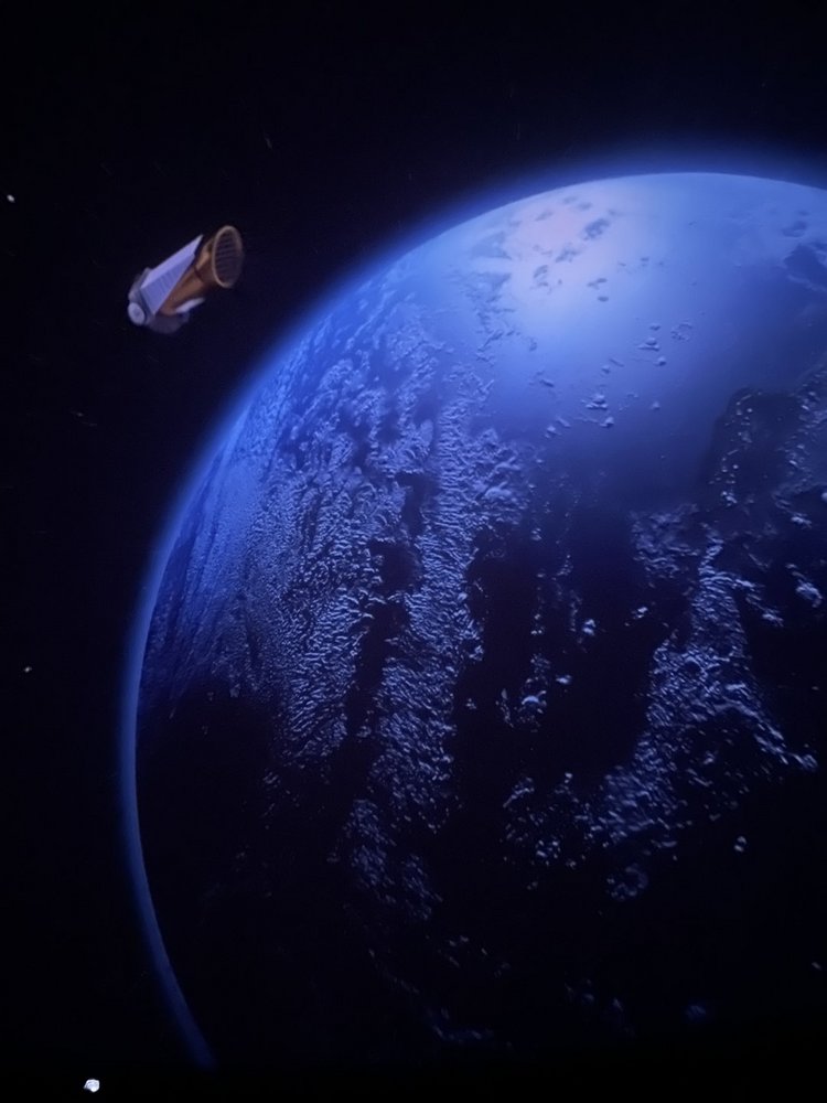 Na zdjęciu widać fragment błękitnej Ziemi oglądanej z przestrzeni kosmicznej. Planeta zajmuje większą część kadru, a jej powierzchnia jest pokryta ciemnymi oceanami i jasnymi, rozciągniętymi chmurami. Po lewej stronie, na tle czarnej przestrzeni kosmosu, unosi się niewielki statek kosmiczny o cylindrycznym kształcie, z jasnym korpusem i panelami przypominającymi panele słoneczne. Całość sprawia wrażenie spokojnej sceny obserwacji Ziemi z orbity.