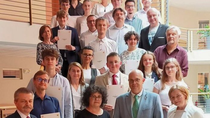 Na zdjęciu znajduje się duża grupa osób, stojących na schodach wewnątrz budynku. Większość z nich trzyma certyfikaty i się uśmiecha. Osoby w grupie są elegancko ubrane. Na pierwszym planie, po prawej stronie, znajduje się mężczyzna w zielonym garniturze oraz kobieta z ciemnymi, kręconymi włosami ubrana na czarno. Wszyscy pozują na tle wnętrza budynku z balustradami i otwartą przestrzenią. 