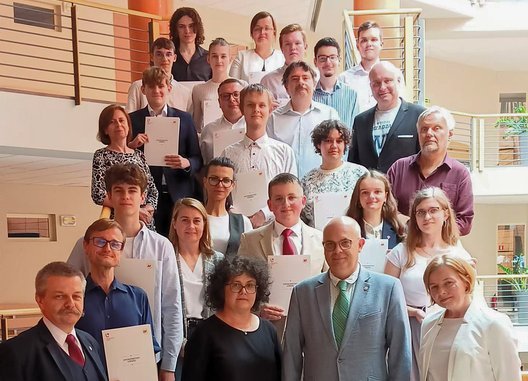 Na zdjęciu znajduje się duża grupa osób, stojących na schodach wewnątrz budynku. Większość z nich trzyma certyfikaty i się uśmiecha. Osoby w grupie są elegancko ubrane. Na pierwszym planie, po prawej stronie, znajduje się mężczyzna w zielonym garniturze oraz kobieta z ciemnymi, kręconymi włosami ubrana na czarno. Wszyscy pozują na tle wnętrza budynku z balustradami i otwartą przestrzenią. 