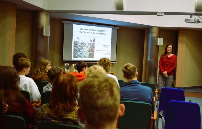 The photo shows a lecture hall. A woman as a lecturer stands in front of the audience.