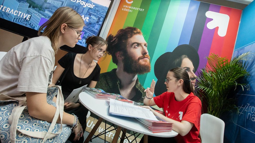 Three young people are sitting at a table and talking; in the background there is a colourful wall displaying the words “University of Lodz”