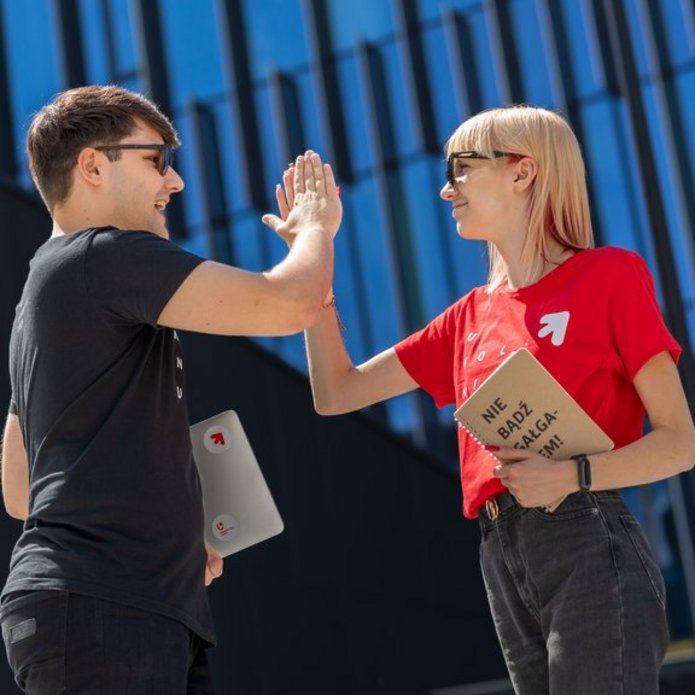 Dwoje młodych ludzi, dziewczyna i chłopak, stoją na dworze w słoneczny dzień i przybijają sobie piątkę