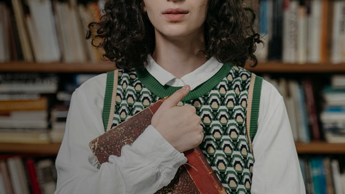 Młoda kobieta o ciemnych, kręconych włosach stoi na tle regału z książkami. Ubrana jest w białą koszulę i zielony sweter w geometryczny wzór. Trzyma w ramionach starą, oprawioną książkę i patrzy spokojnie w obiektyw.