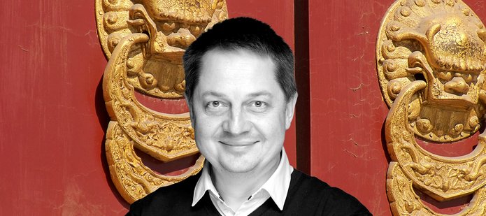 Czarno-biały wizerunek profesora Mierzejewskigo w czarnym swetrze z widocznymi spod niego białymi kołnierzykami, w tle dwie duże złote, azjatyckie płaskożeźby w kształcie kołatek do drzwi