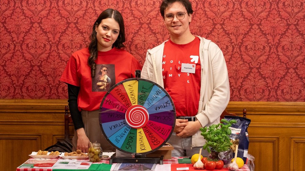 Dziewczyna i chłopak stoją uśmiechnięci przy stole na tle czerwonej, ozdobnej ściany. 