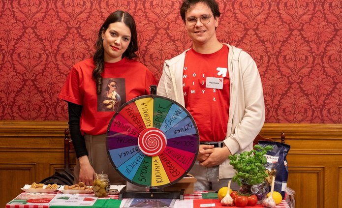 Dziewczyna i chłopak stoją uśmiechnięci przy stole na tle czerwonej, ozdobnej ściany. 