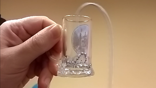 Na zdjęciu widać dłoń trzymającą niewielkie, przezroczyste szklane naczynie przypominające mały kieliszek. Wewnątrz naczynia znajdują się drobne, metaliczne kulki. Z prawej strony do naczynia wprowadzona jest przezroczysta, cienka rurka, która wygina się łukiem nad jego krawędzią i prowadzi na zewnątrz. Tło zdjęcia jest jednolite i jasne, w kolorze beżowym, dzięki czemu główny element – szklane naczynie z kulkami i rurką – jest dobrze widoczny na pierwszym planie. Całość wygląda jak fragment prostego doświadczenia fizycznego z użyciem małego naczynia, kulek i elastycznej rurki.