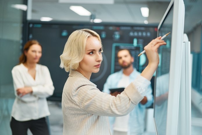 na obrazku: młoda kobieta, krótkie blond włosy, jasna marynarka, stoi bokiem i zapisuje coś na tavlicy. Dalej w tle widoczna jest młoda kobieta i młody mężczyzna, którzy obserwują kobietę przy tablicy