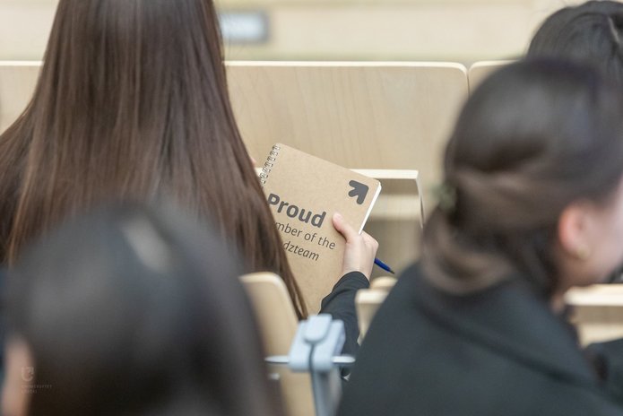 Na zdjęciu widać grupę osób siedzących tyłem do nas. Jedna z nich trzyma w dłoniach notes w kolorze beżowym, na którym jest napis proud member of the unilodz team